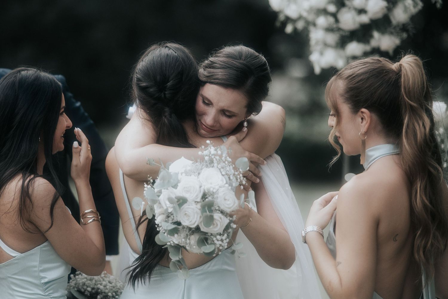 Bridesmaids in dark dresses share emotional moment with bride holding white bouquet.