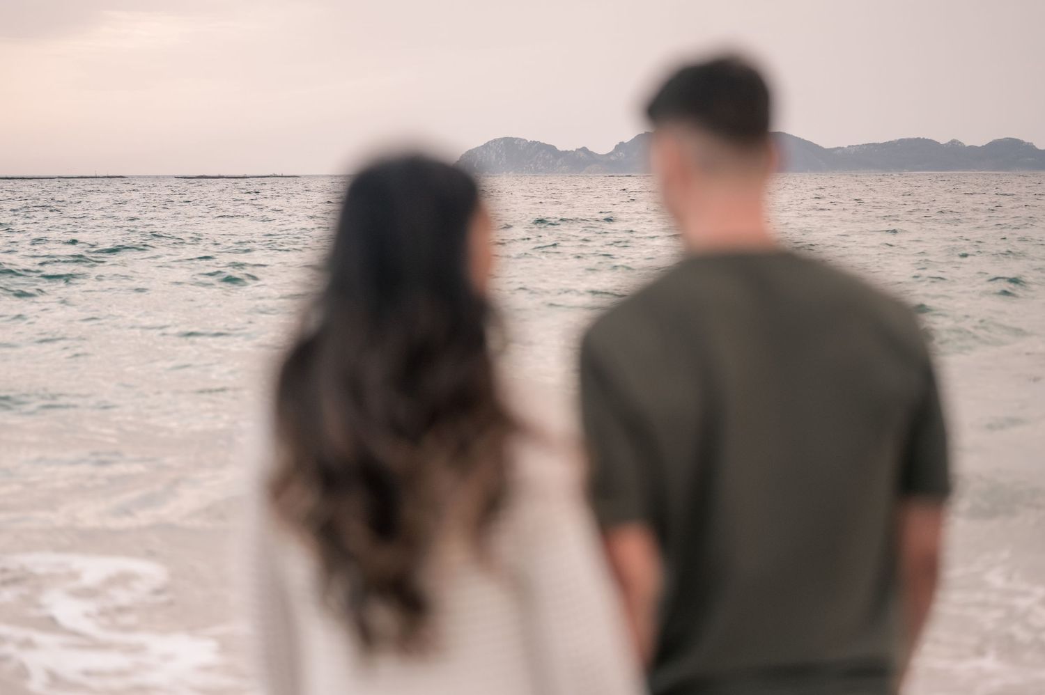 Couple walking along a misty beach at sunset holding hands with mountains in the background.