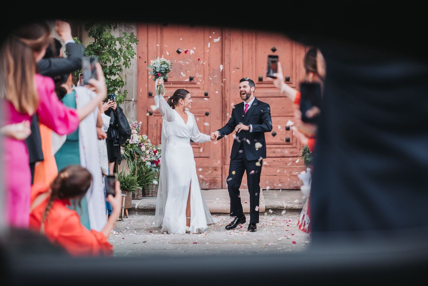 Wedding party celebrates joyfully outside a rustic brick building during an outdoor ceremony.