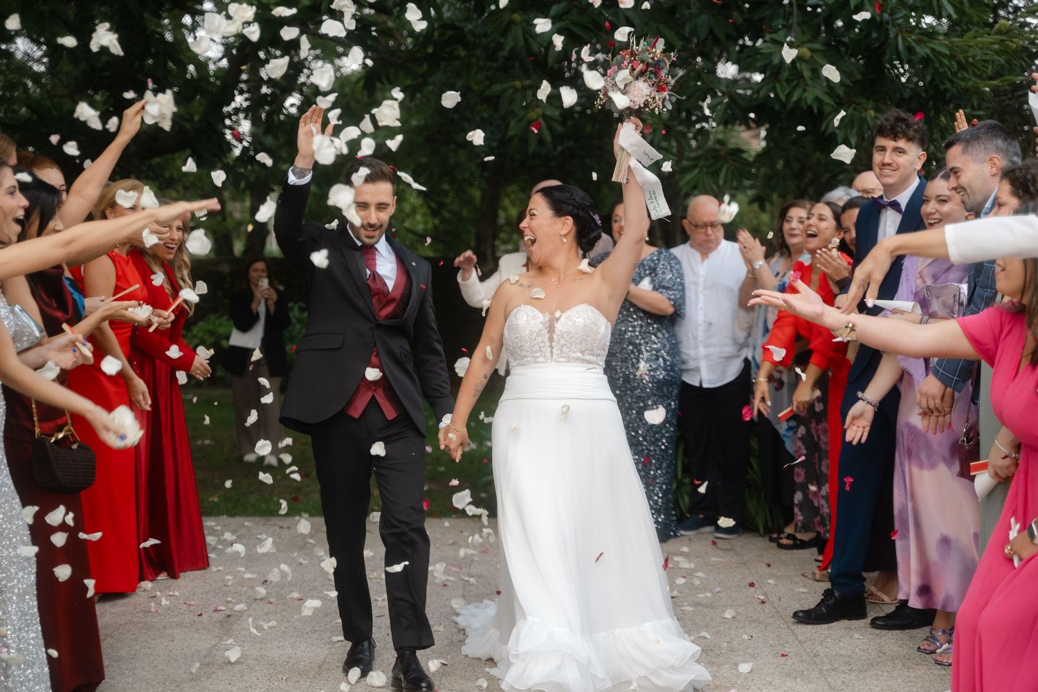 Wedding guests shower a couple with white flower petals during a joyous outdoor celebration.