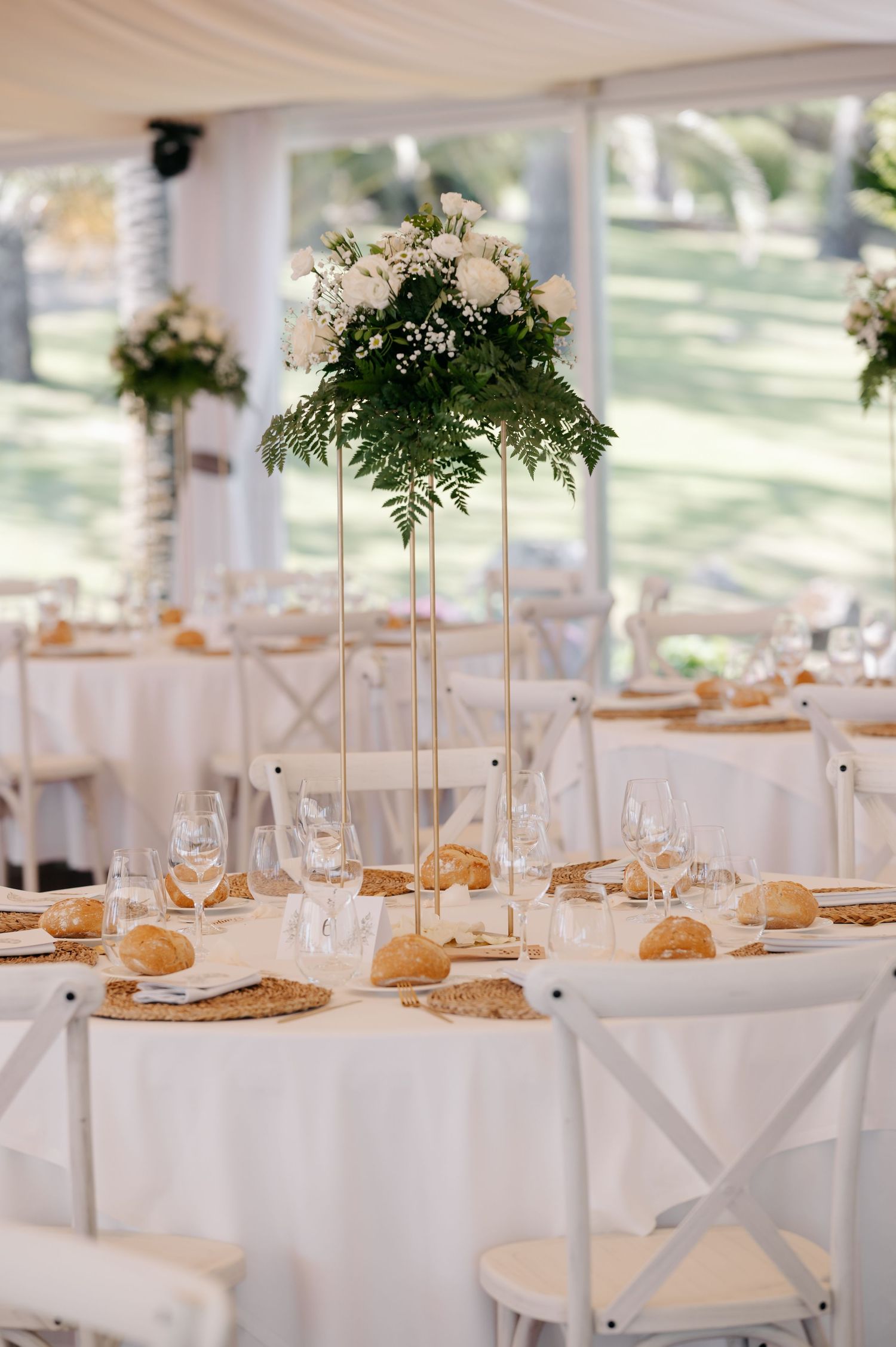 Elegant wedding reception table with tall white floral centerpiece, white linens, and rustic bread rolls on round tables.