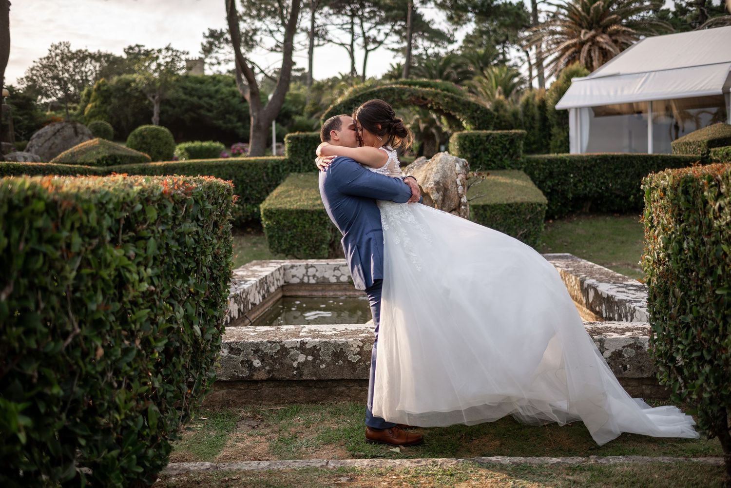 A romantic wedding moment in a garden with manicured hedges and palm trees at sunset.