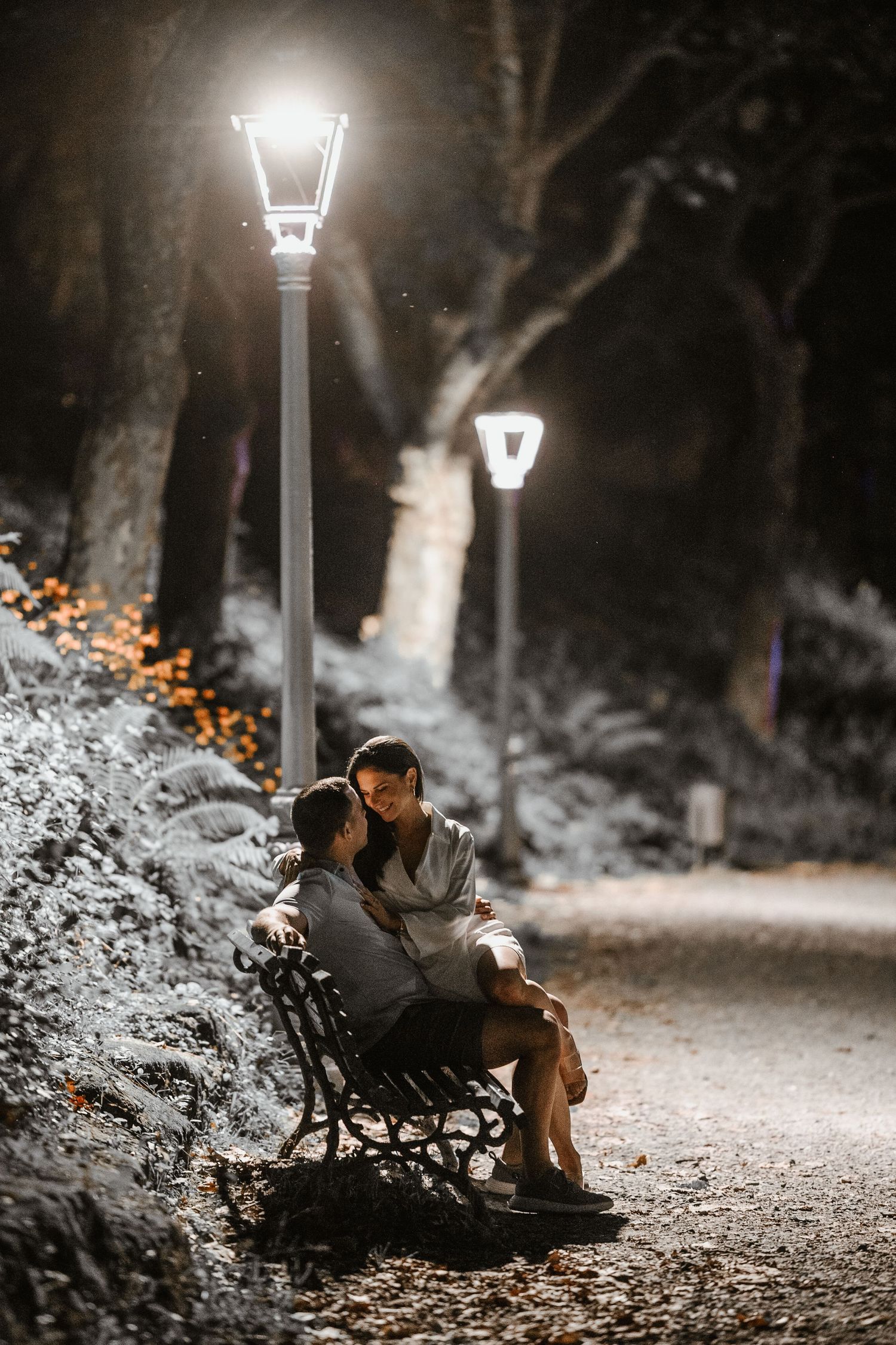 A couple sits on a park bench at night under illuminated lamp posts with snow-covered trees in the background.