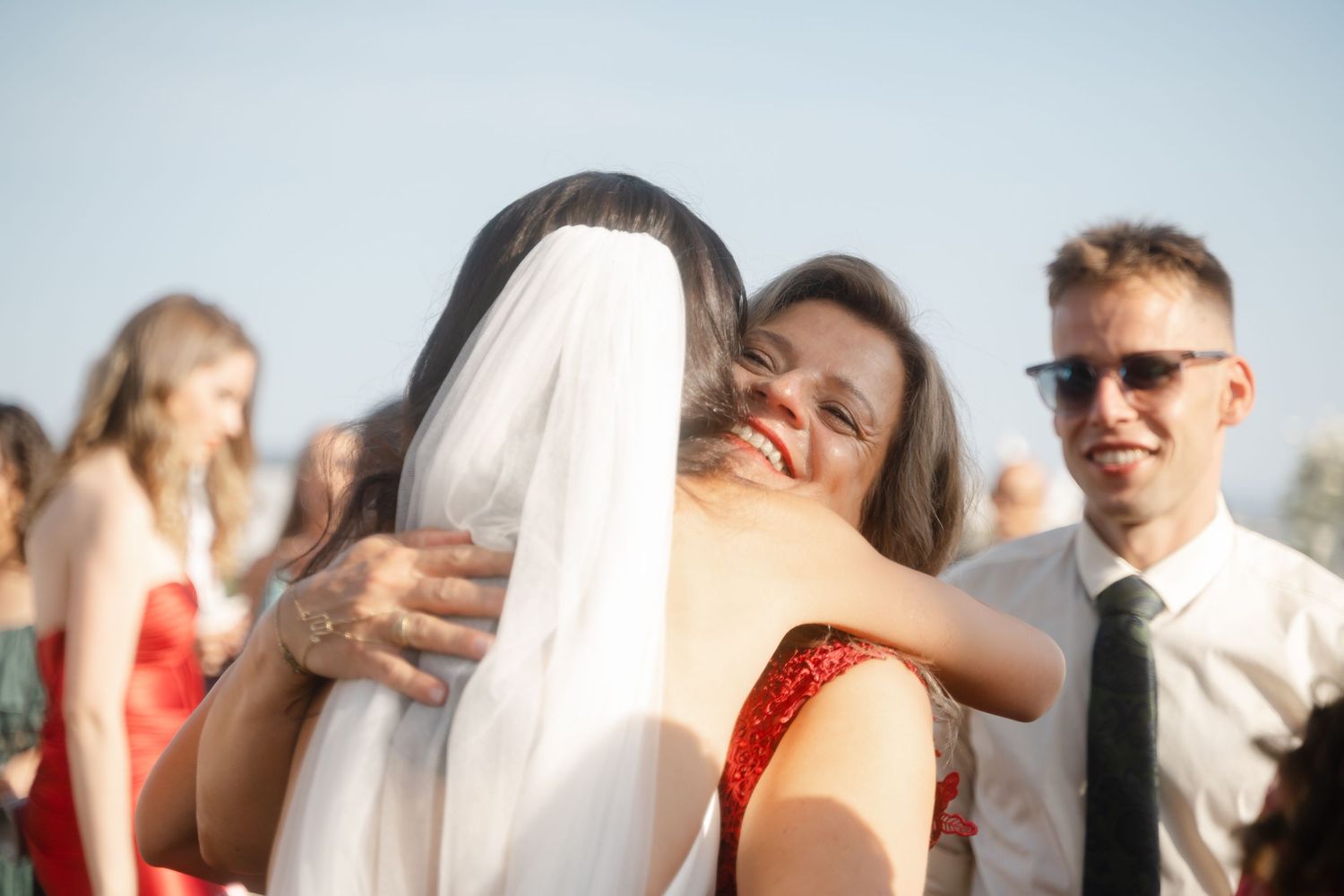 Wedding guests embrace and celebrate during an outdoor reception under sunny skies.