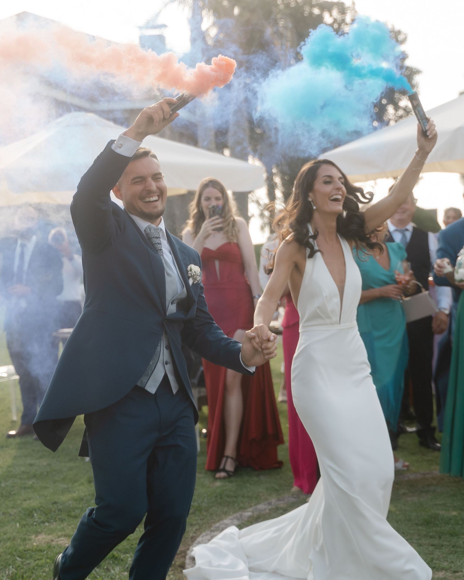 Wedding celebration with colorful smoke bombs creating vibrant blue clouds as guests dance and celebrate outdoors.