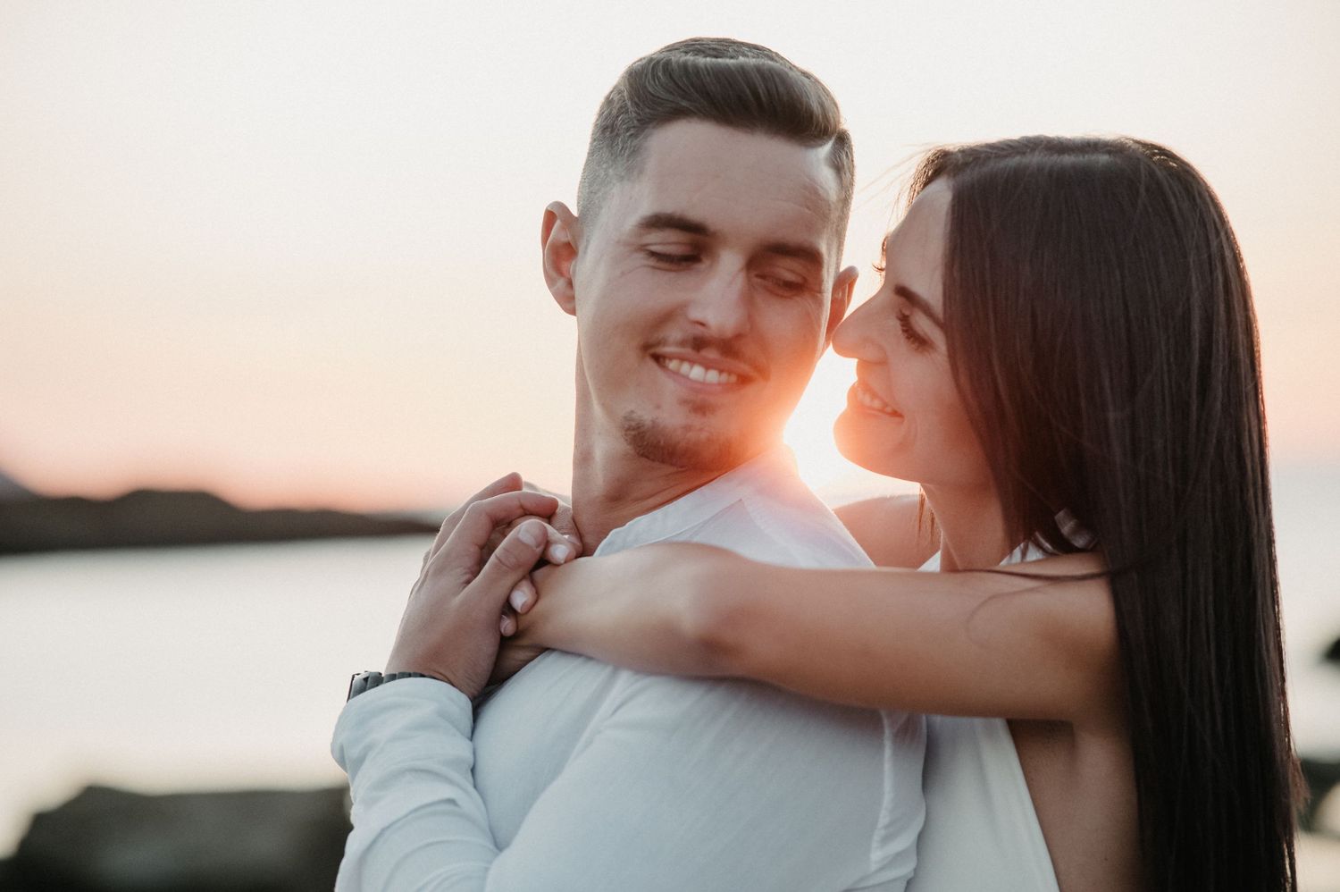 Romantic couple embraces during sunset photoshoot by the water in matching white outfits.