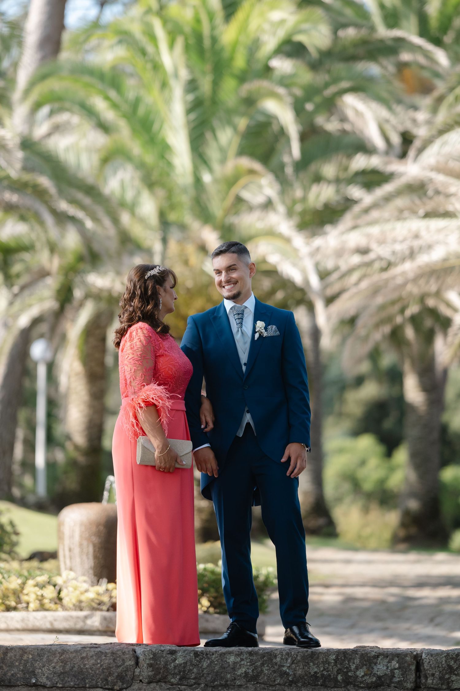A couple shares a romantic moment while walking along a palm tree-lined path in formal attire.