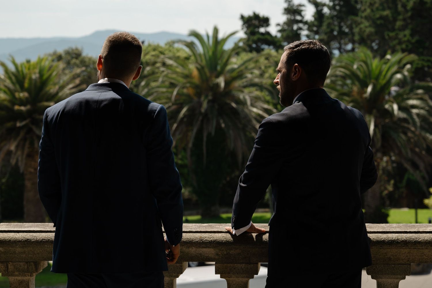 Two people in dark suits stand on a terrace overlooking palm trees in a scenic outdoor setting.