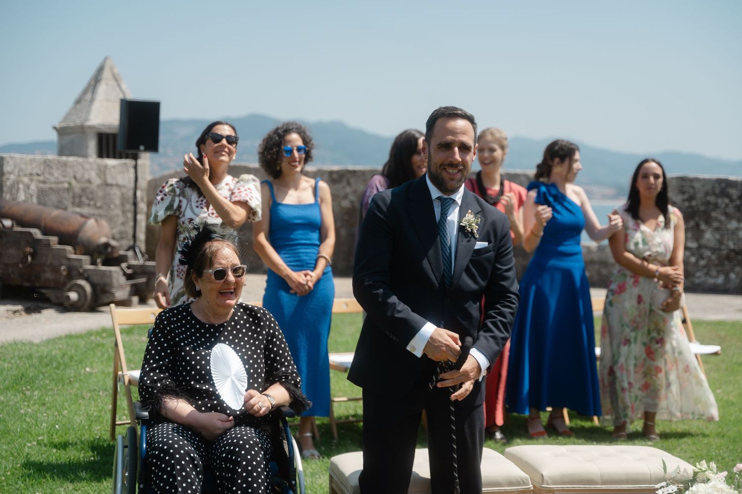 A group of wedding guests gather outdoors on a sunny day near a stone structure.