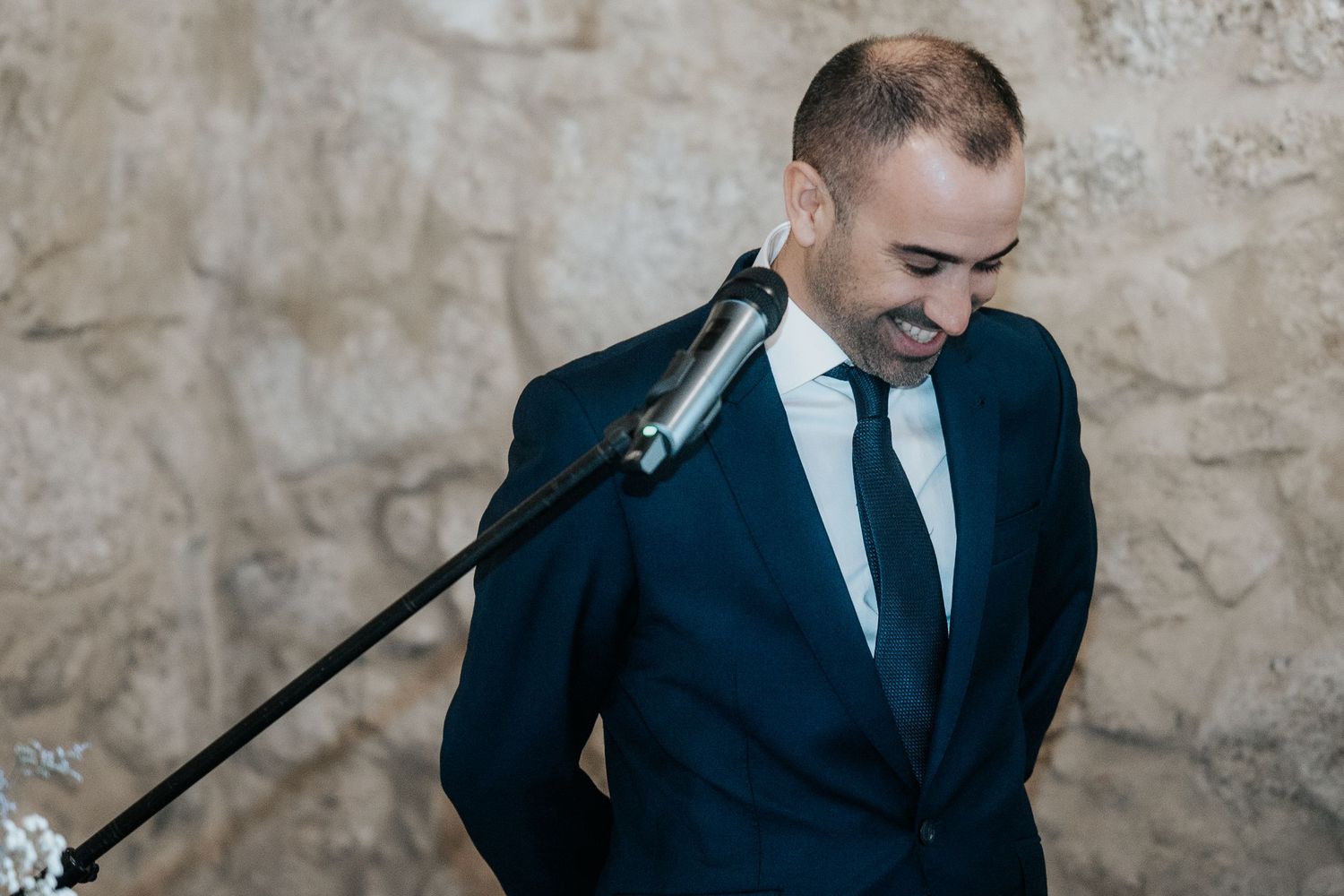 A person in a navy suit plays an acoustic guitar while seated against a rustic stone wall background.