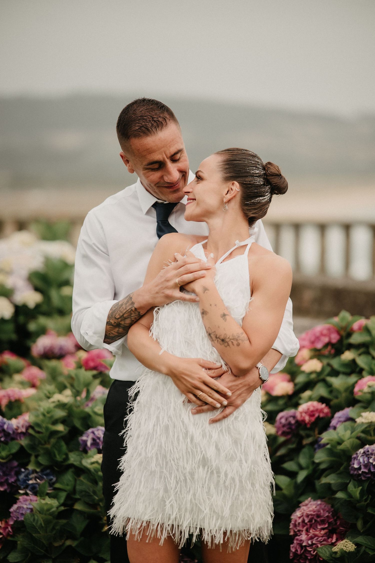 Romantic couple in wedding attire embrace near blooming hydrangea flowers during sunset by the ocean.