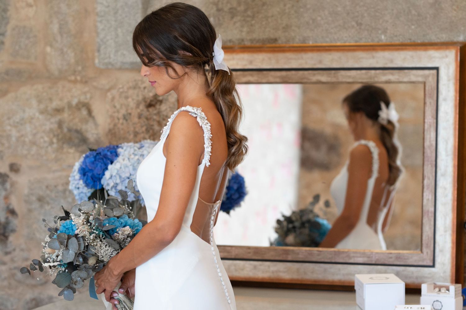 A bride in a white wedding dress holds a blue and white bouquet while looking in a mirror.