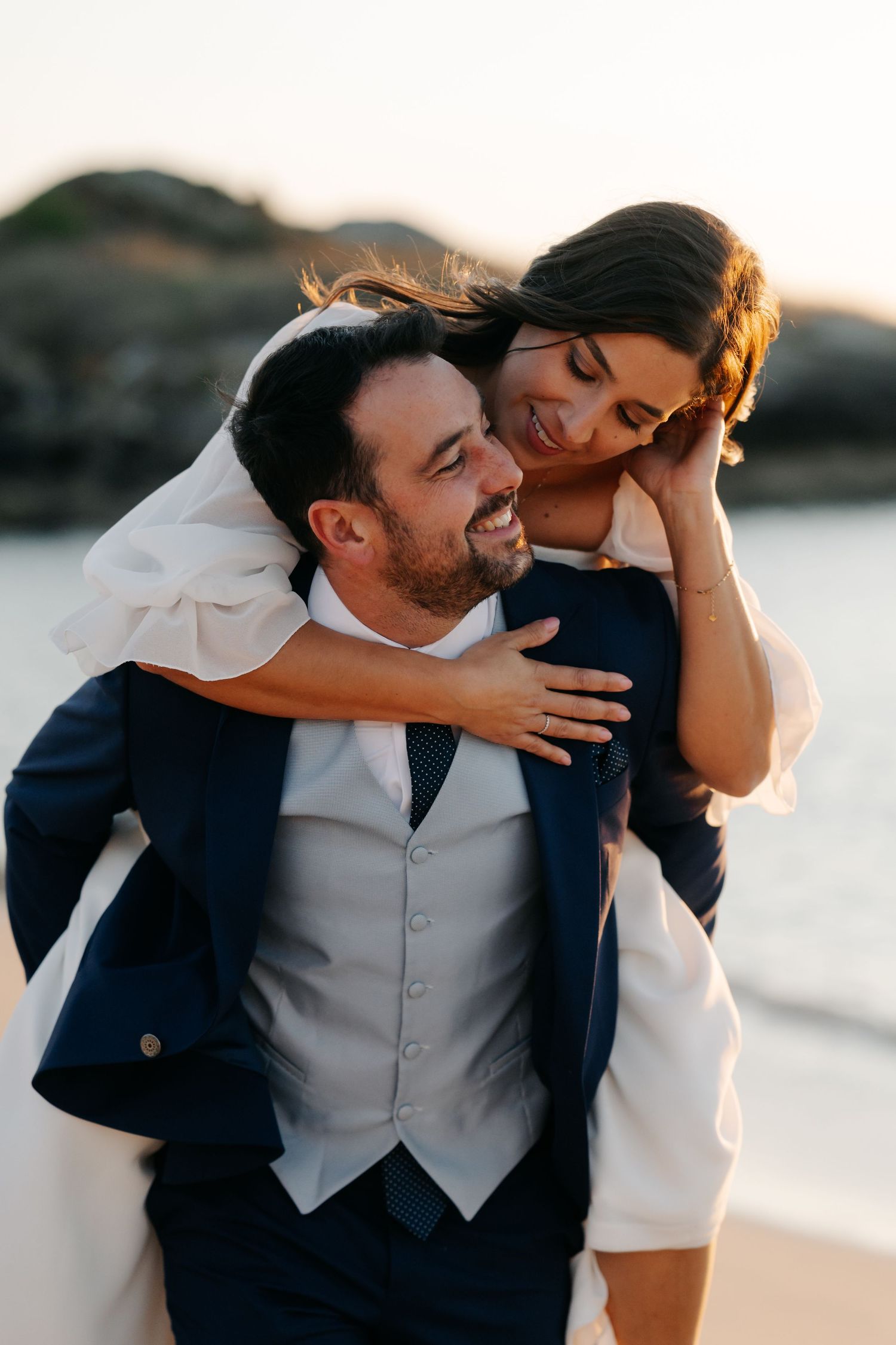 A joyful couple shares a tender moment on the beach at sunset, the bride playfully embracing from behind.