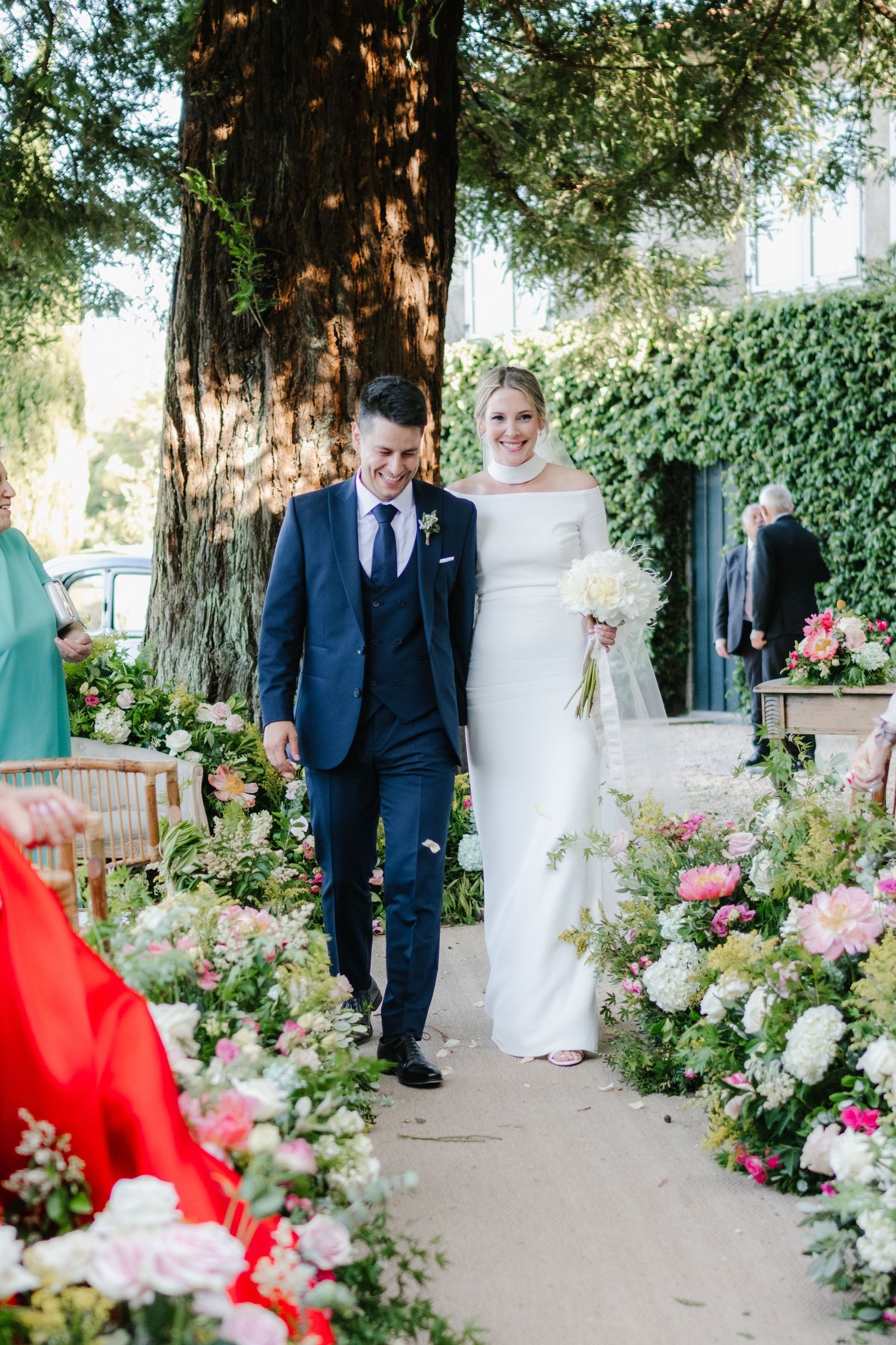 A wedding couple walks through a garden pathway decorated with lush pink and white floral arrangements.