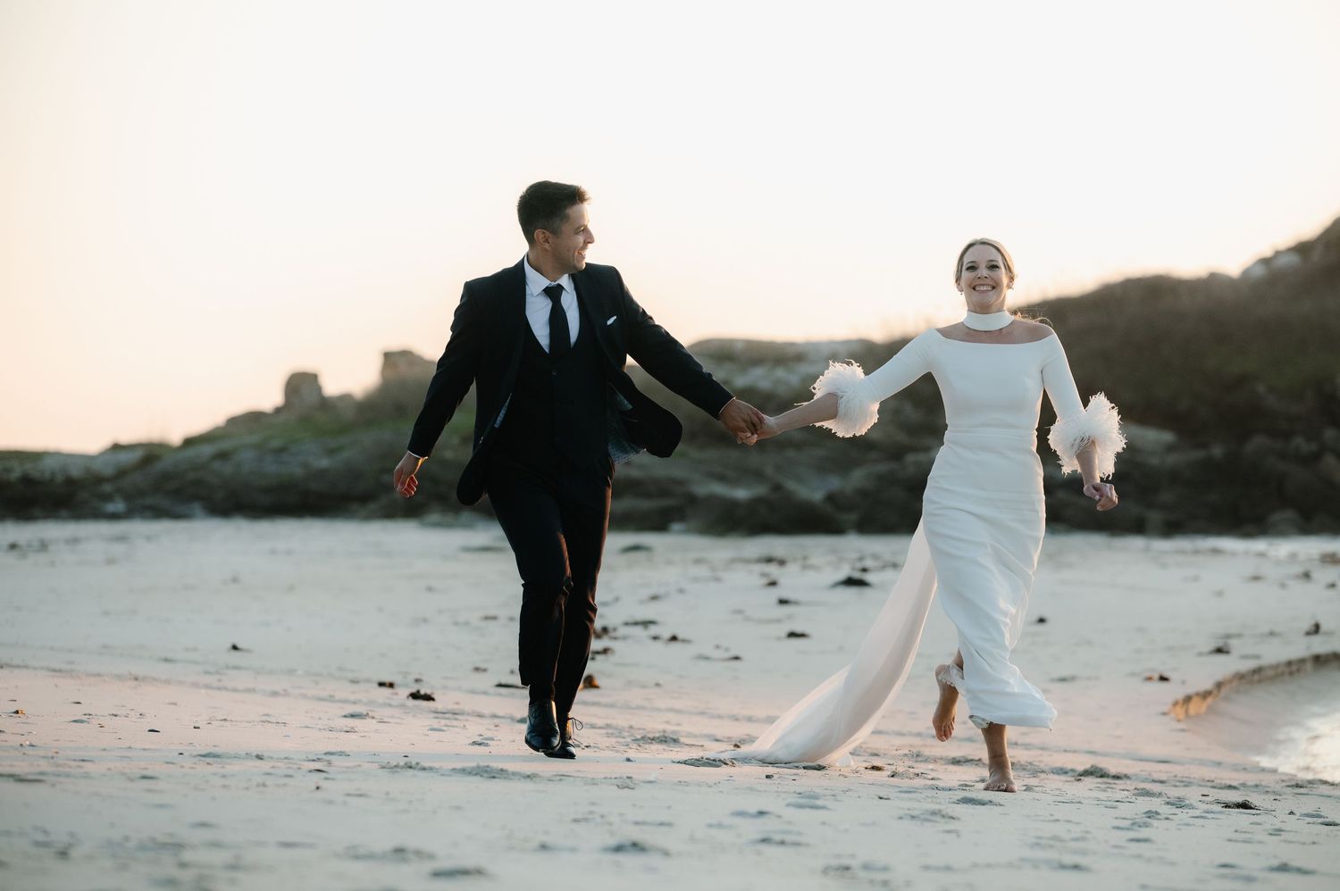 Couple walks along a scenic beach at sunset with flowing white dress trailing behind.
