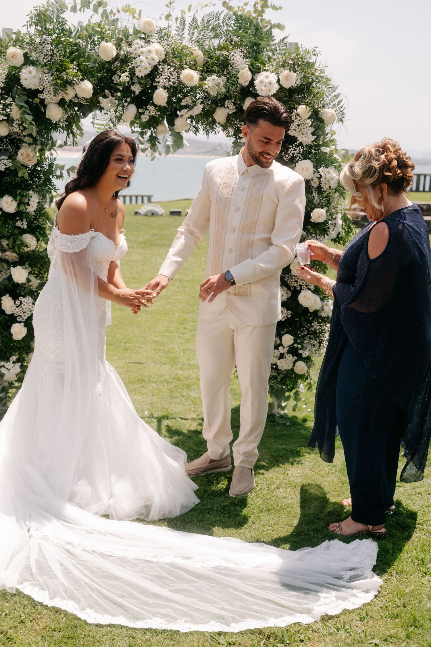 A wedding couple dances joyfully under a floral arch decorated with white roses and greenery on a sunny lawn.