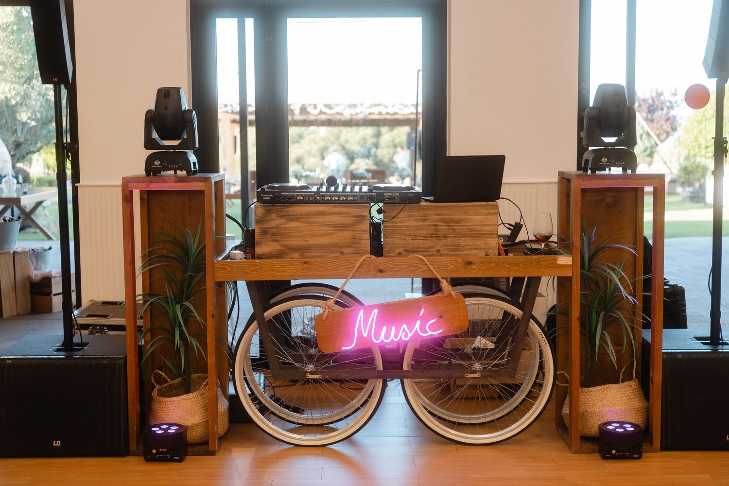 A wooden DJ booth setup with neon music sign and speakers against a bright window backdrop.
