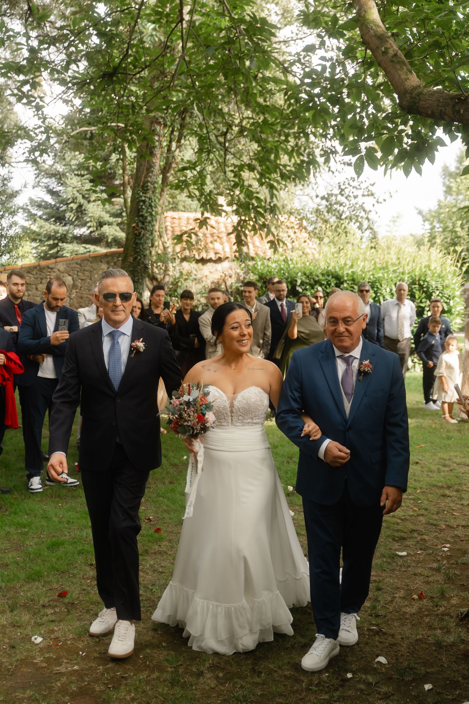 Wedding guests gather outdoors under trees during a summer celebration.