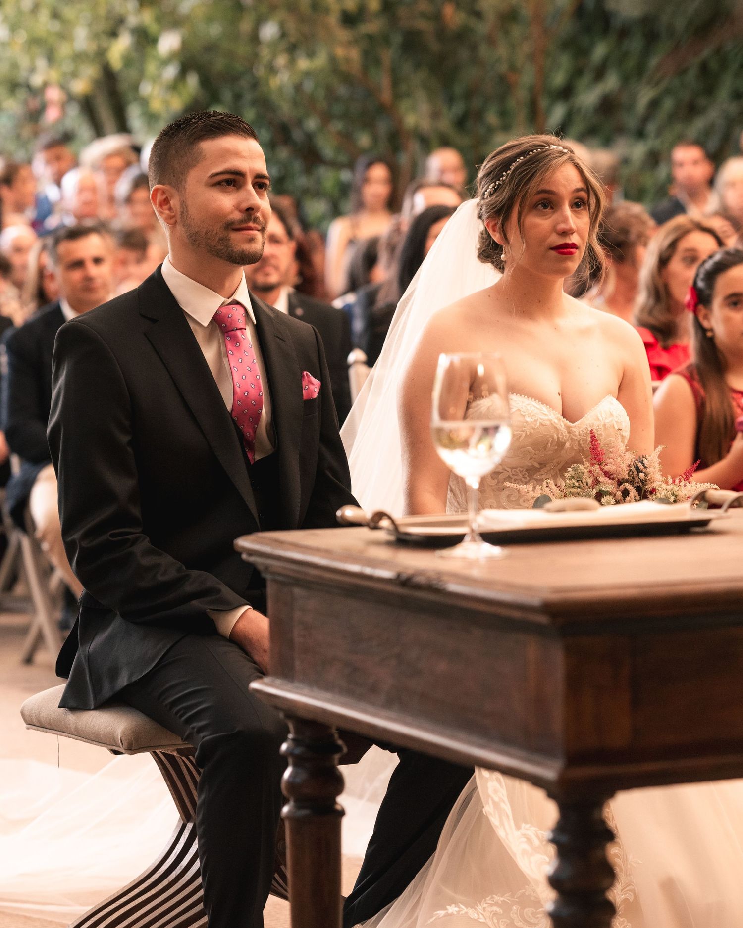 A bride and groom sit together at a wooden table during their wedding ceremony with guests in the background.