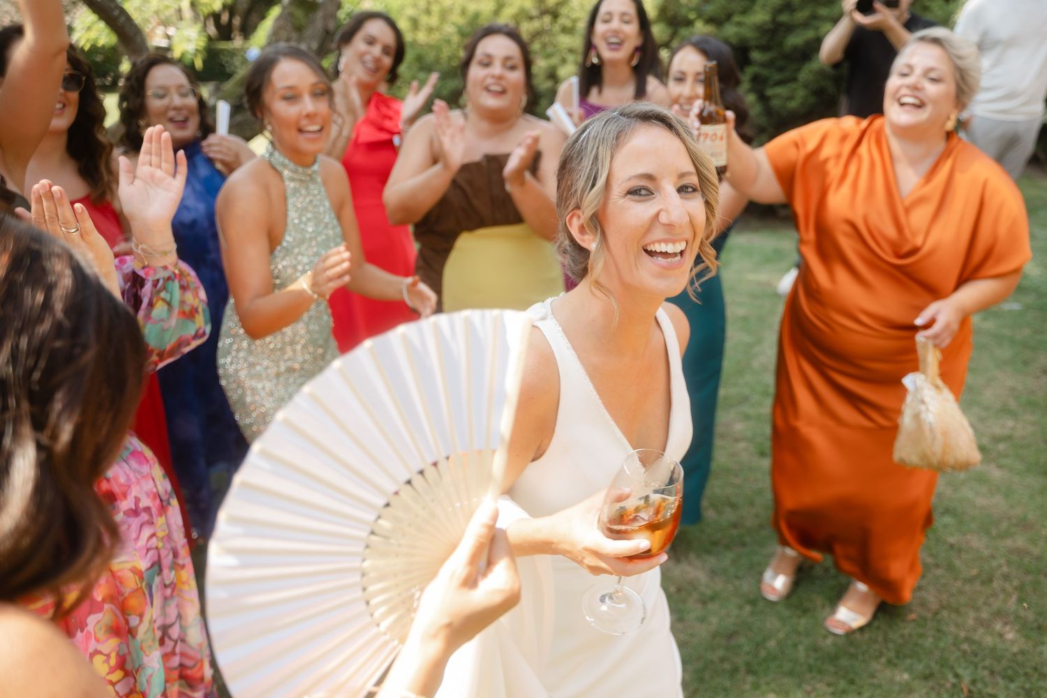 Wedding guests dance and laugh while holding decorative fans at an outdoor celebration.