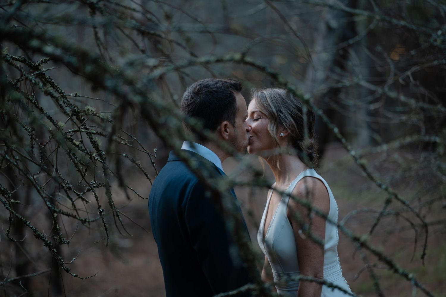 A couple in formal attire share a romantic moment outdoors in a moody, dark setting.