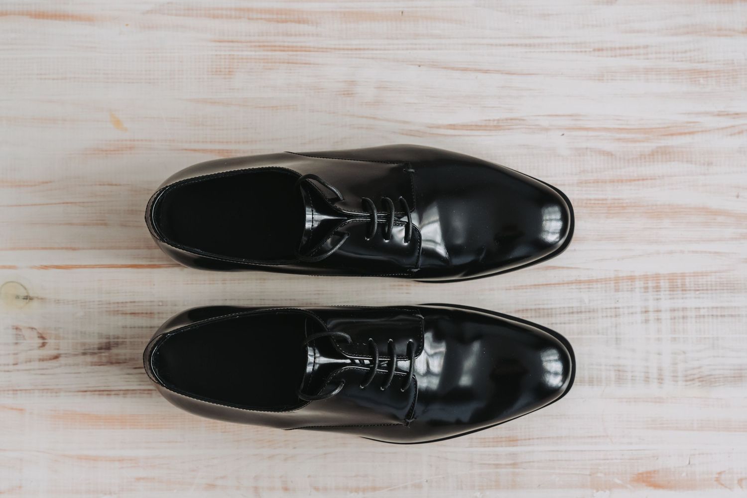 A pair of shiny black leather dress shoes photographed from different angles on a white wooden surface.