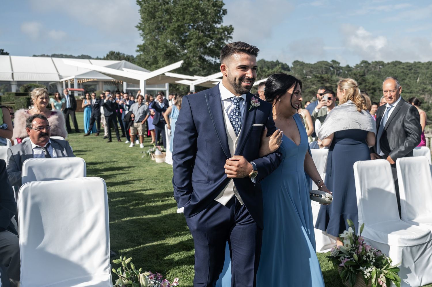 Guests mingle outdoors at an elegant wedding reception with white tents and green lawn in the background.