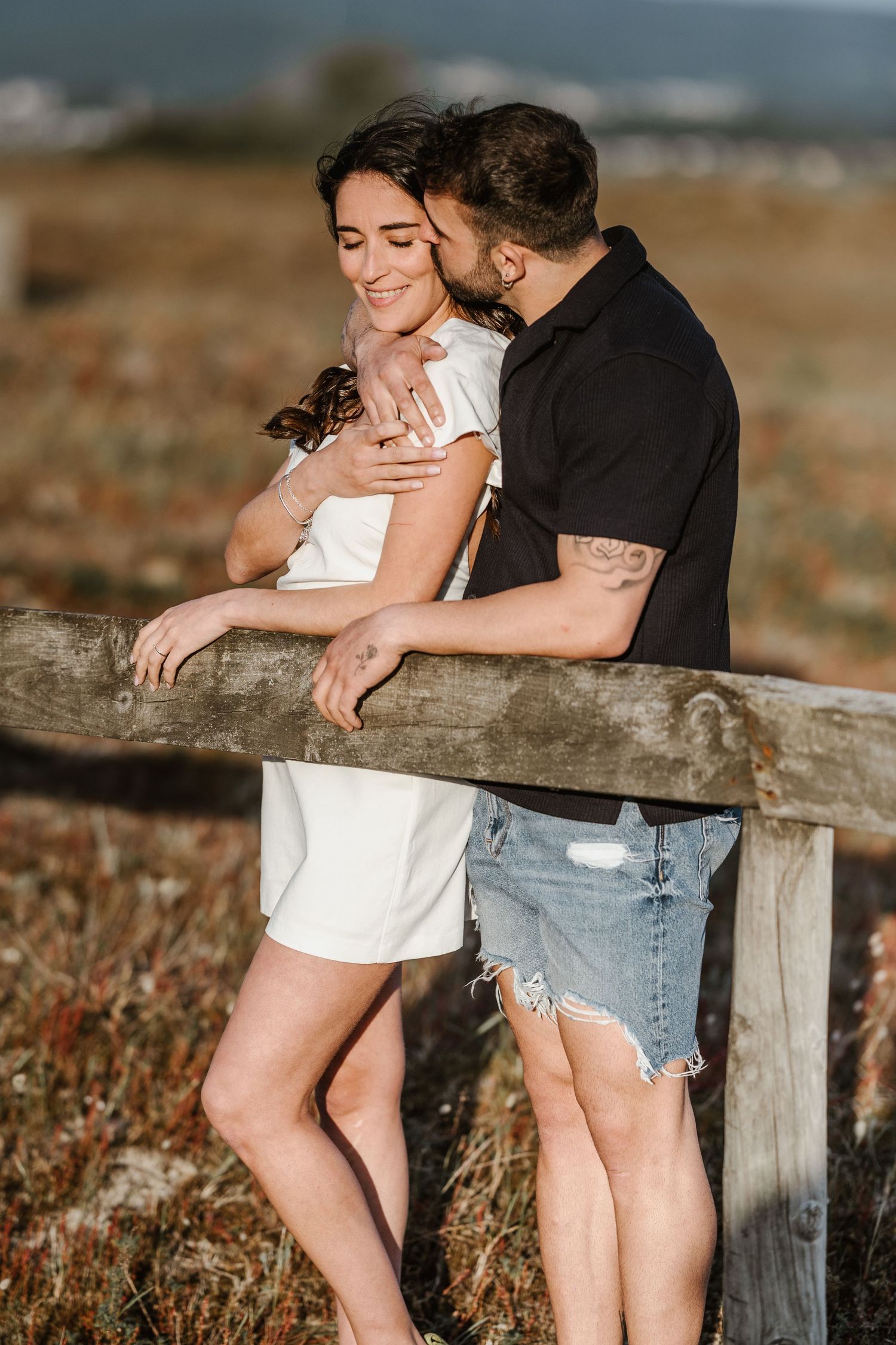 Couple embracing on a rustic wooden fence in a sunlit field during golden hour.