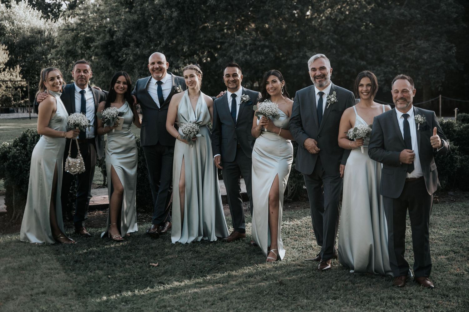 A wedding party poses together outdoors wearing navy suits and light gray dresses with natural greenery in background.