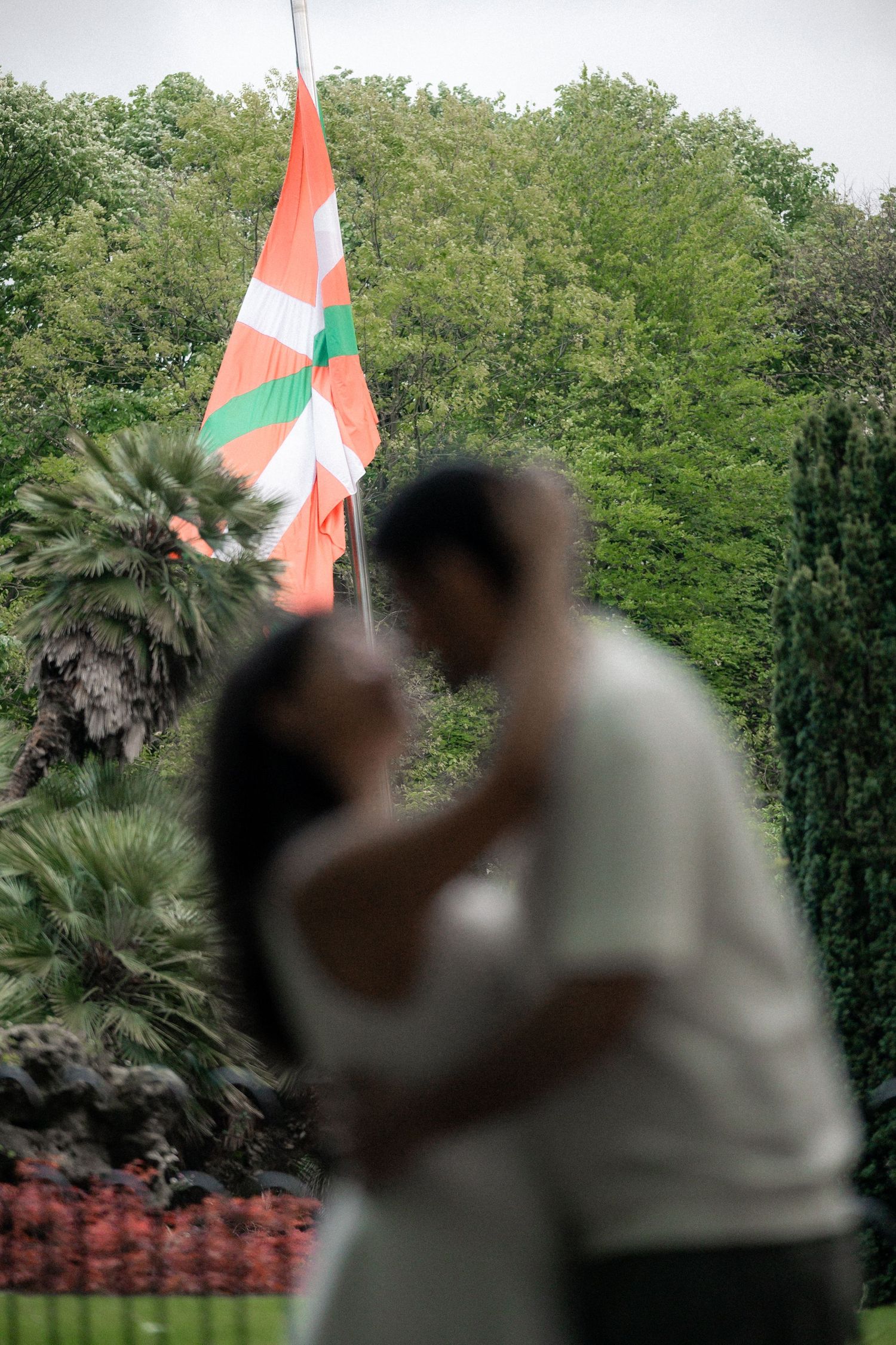 Couple embracing in a lush garden setting with palm trees and an orange and green flag visible.