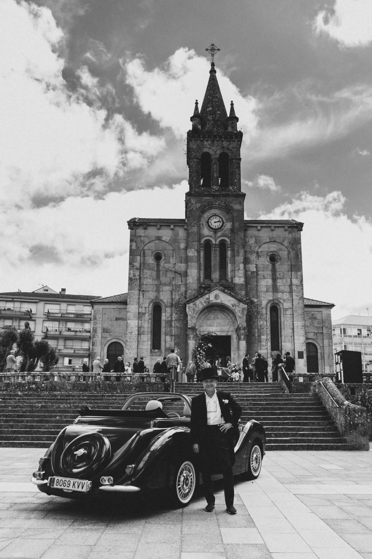 Classic black vintage car parked in front of a grand stone church with Gothic architecture and steps in black and white.