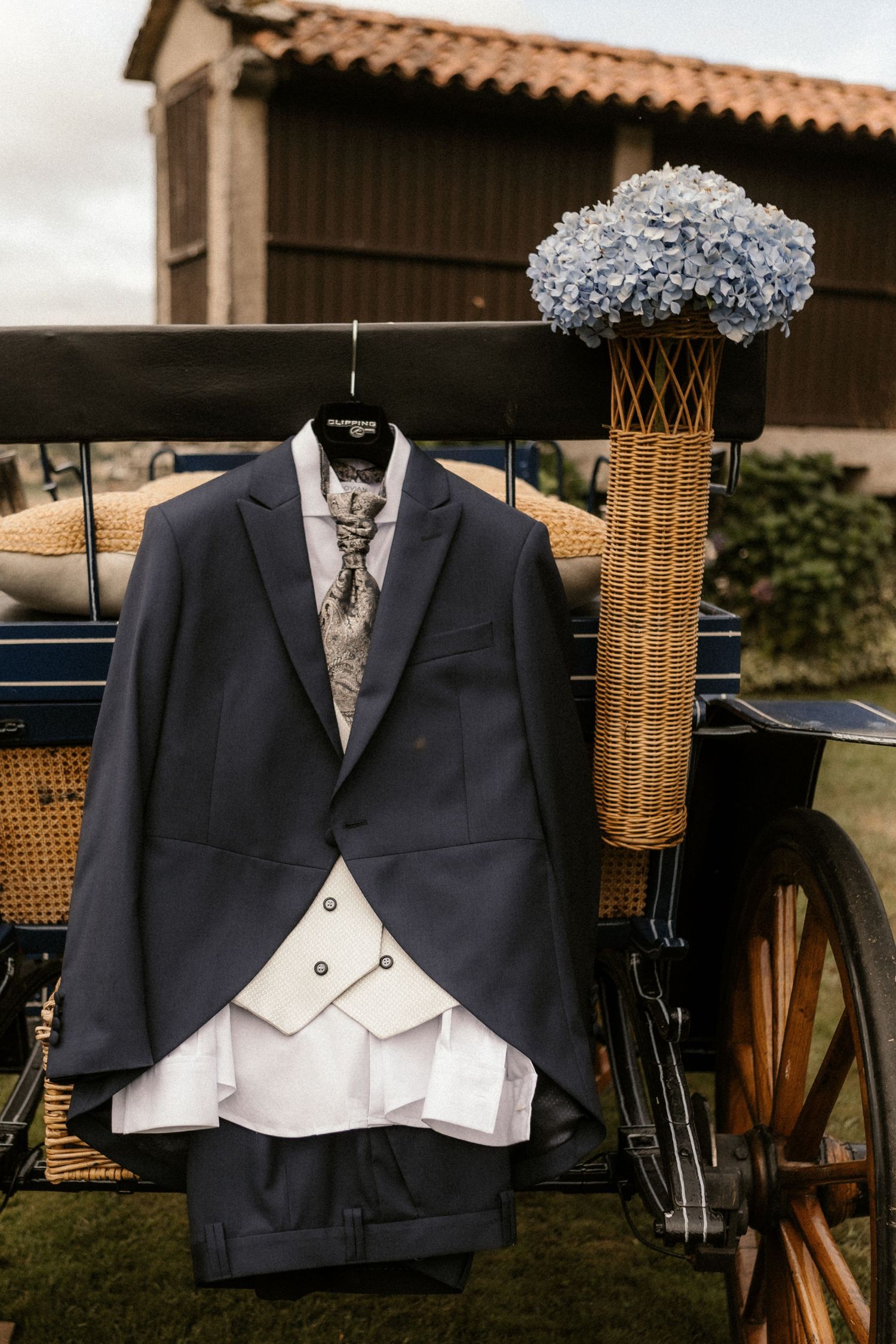 Elegant formal wedding attire with black tuxedo jacket and white vest displayed next to blue hydrangeas in wicker vase.