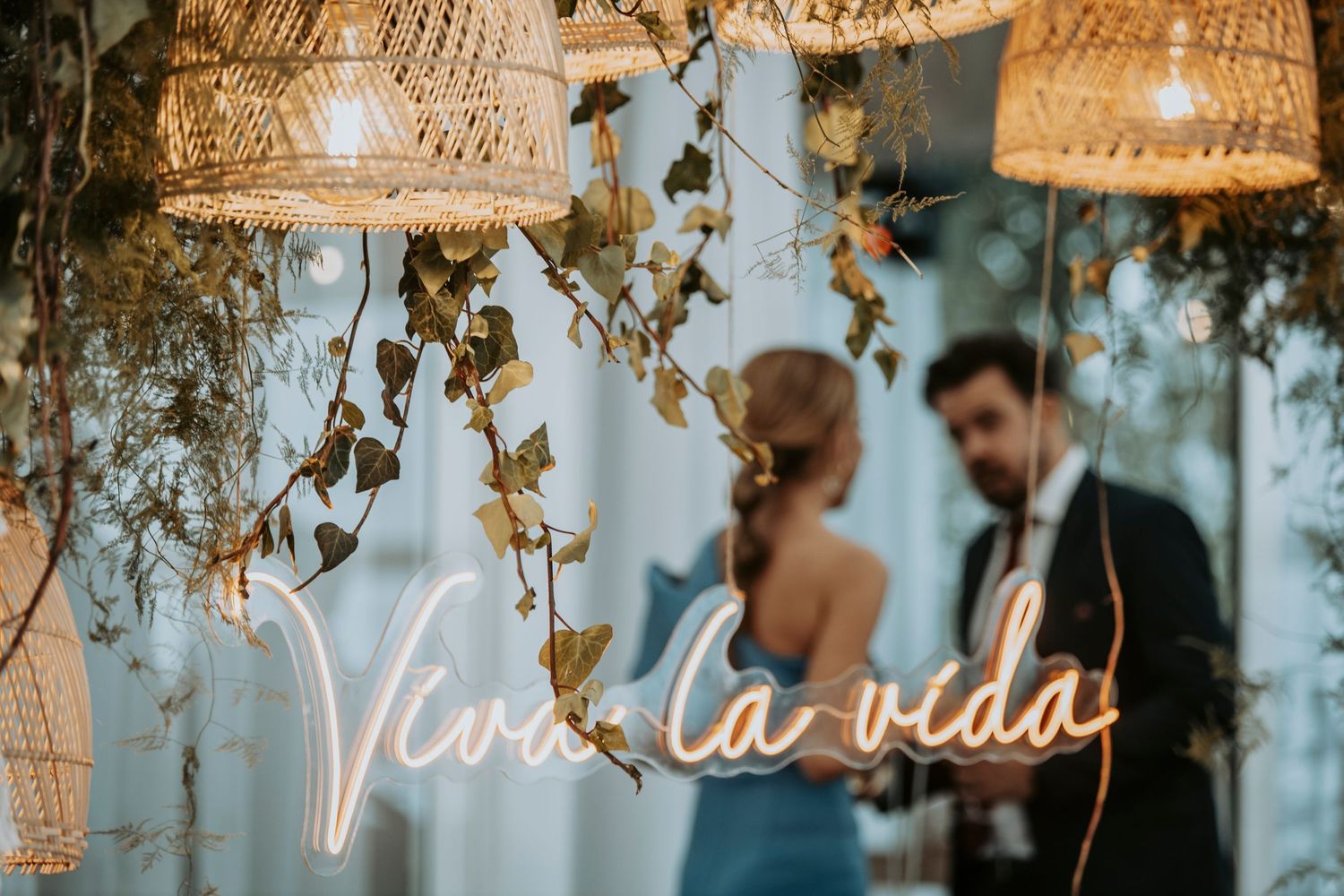 Romantic wedding ceremony setup with woven pendant lamps and neon 'Viva la vida' sign glowing against blue backdrop.
