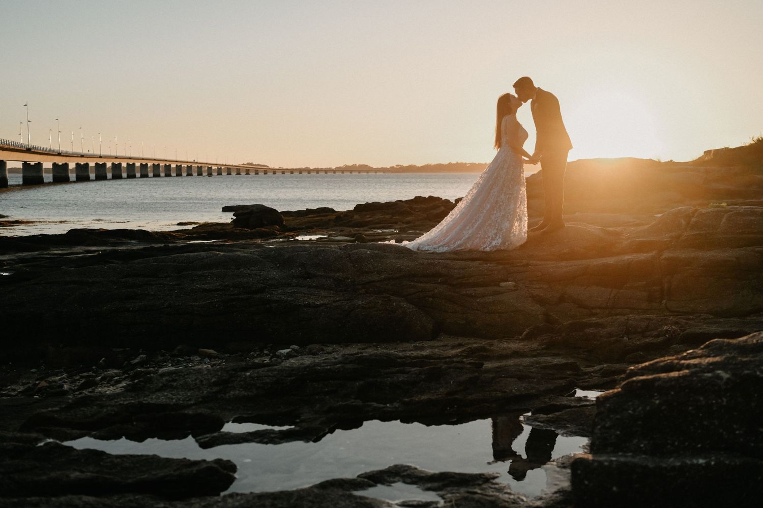 Silhouetted couple embraces on rocky beach during golden sunset with pier in background.