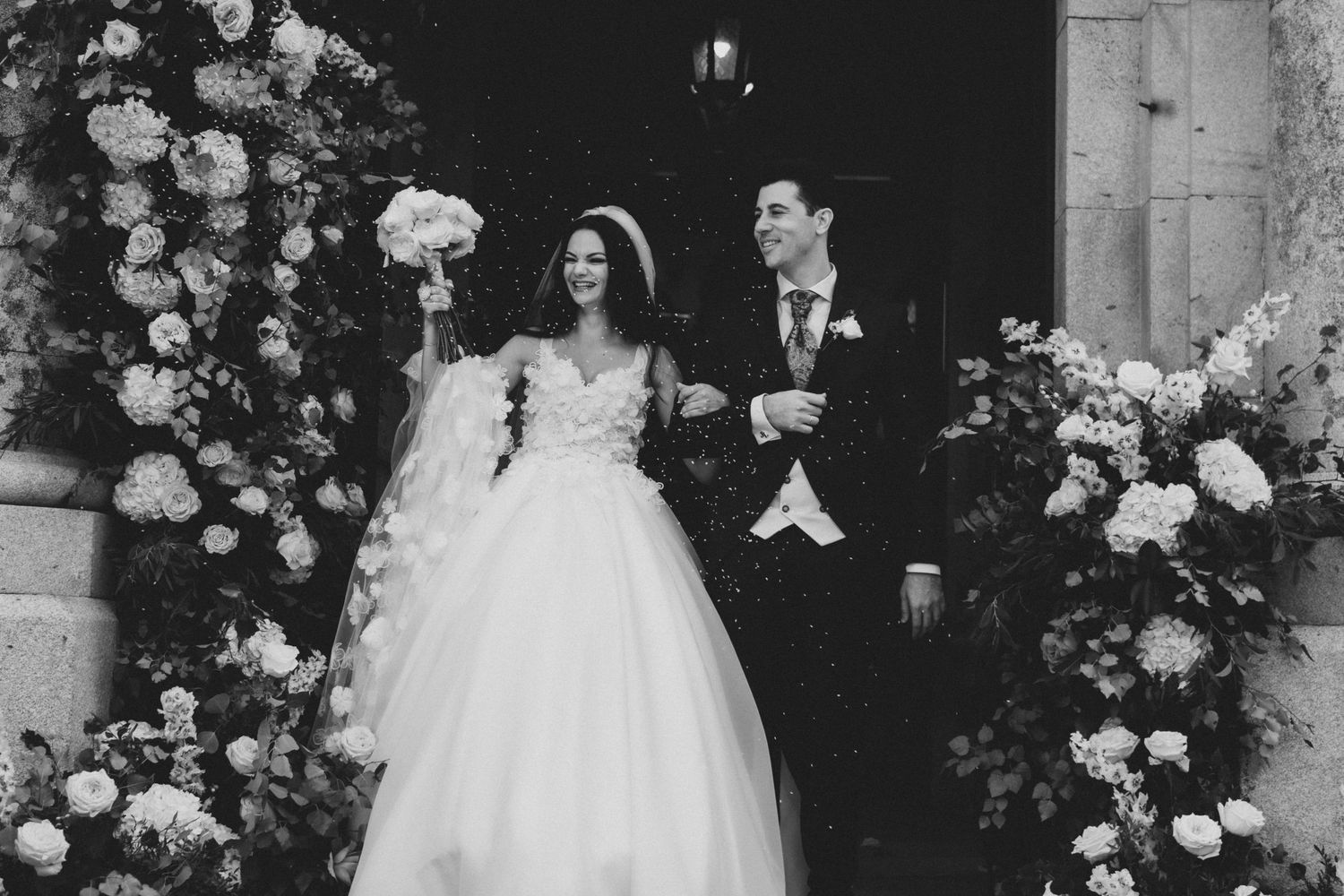 Black and white wedding photo showing couple exiting church with elegant floral arch of white roses and decorations.