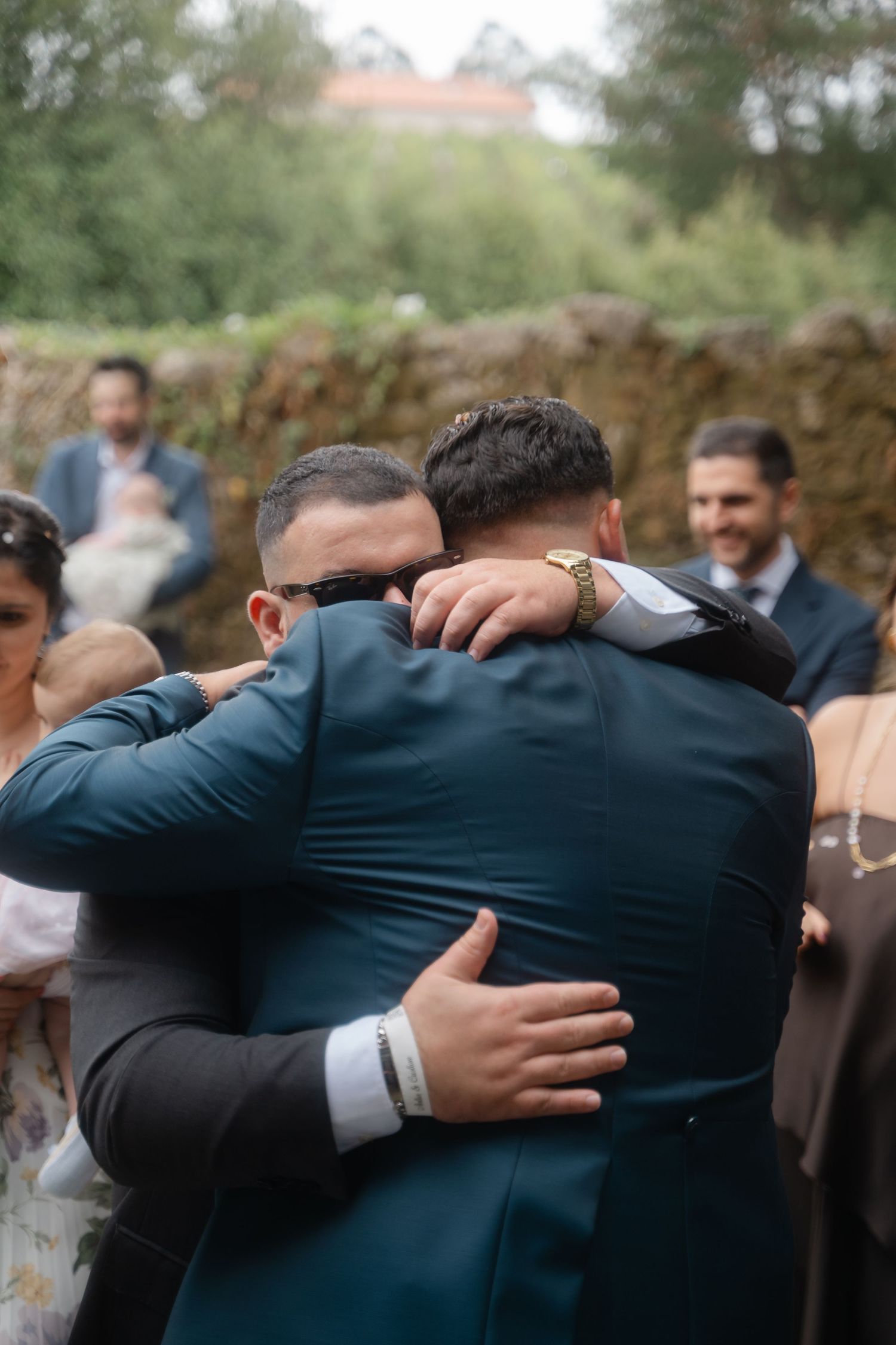 A group of men in dark suits embrace in a supportive huddle at an outdoor gathering.