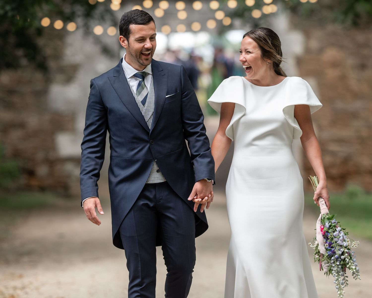 A couple in wedding attire walks and laughs together in front of a stone wall with twinkling lights overhead.