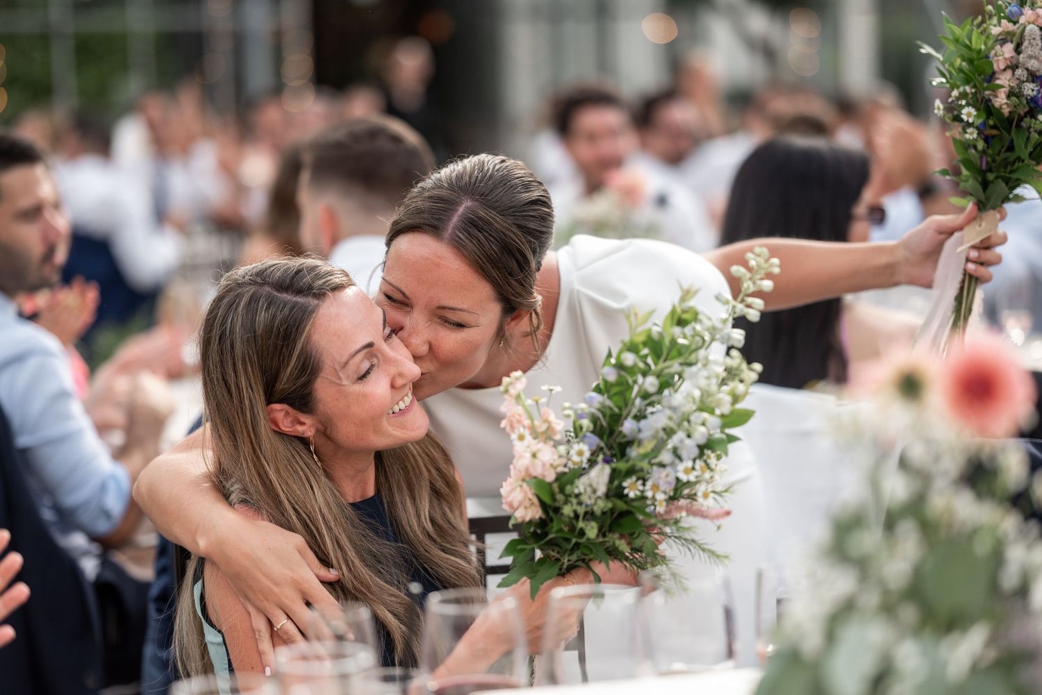 Wedding guests gather at an outdoor reception venue with decorative floral arrangements on tables.