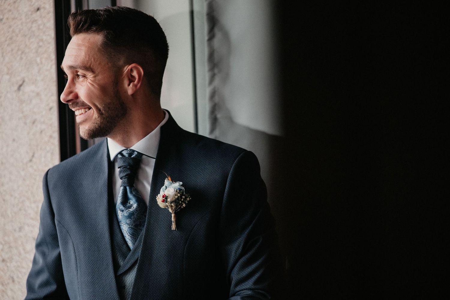 A groom in a navy blue suit and tie smiles while standing against a textured wall in natural light.
