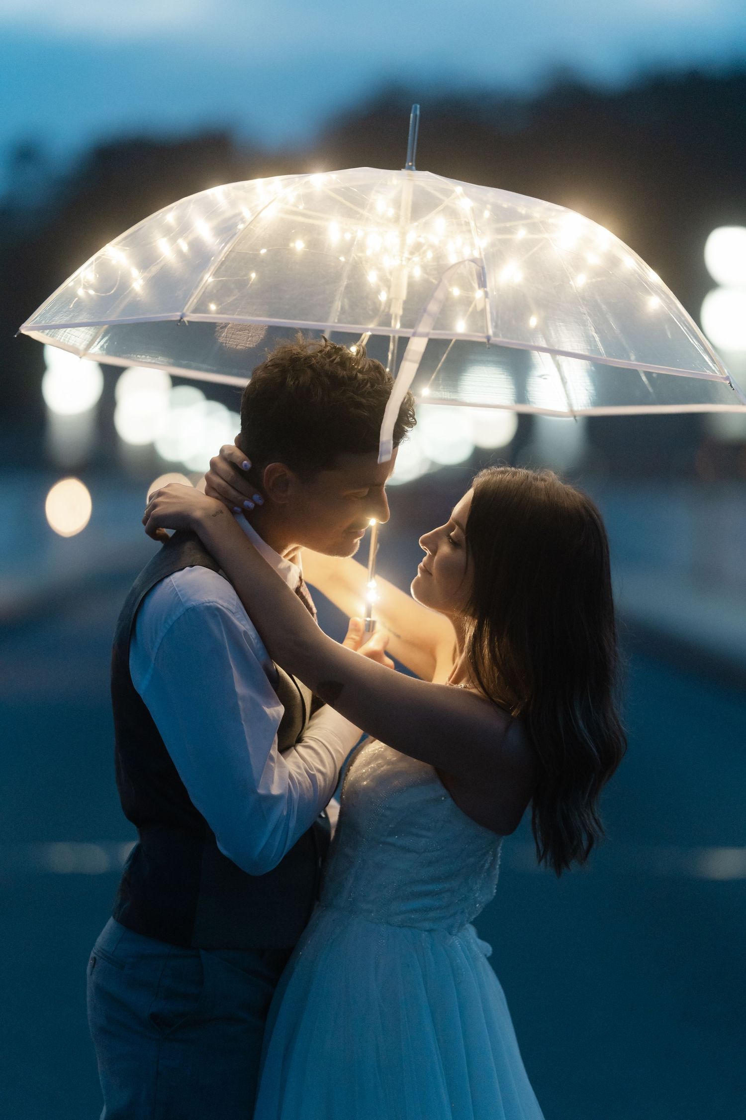 Bride and groom embracing under a glowing fairy light umbrella in the evening.