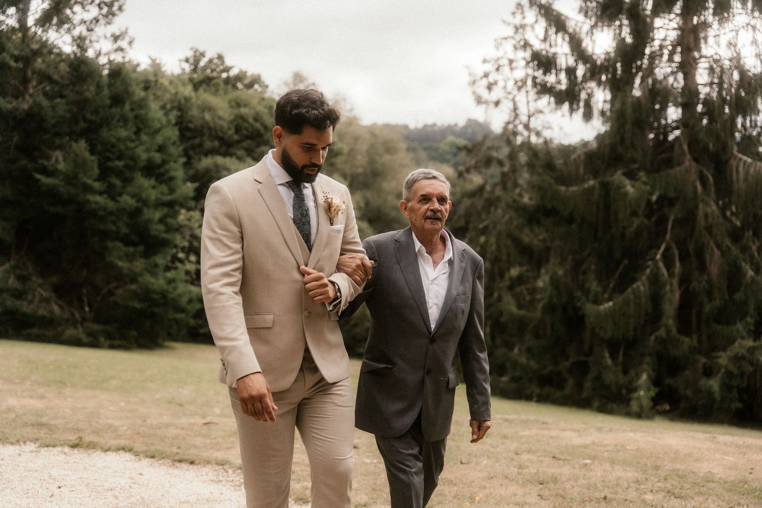 Two people in formal attire walk and talk together in an outdoor setting with trees and natural scenery.