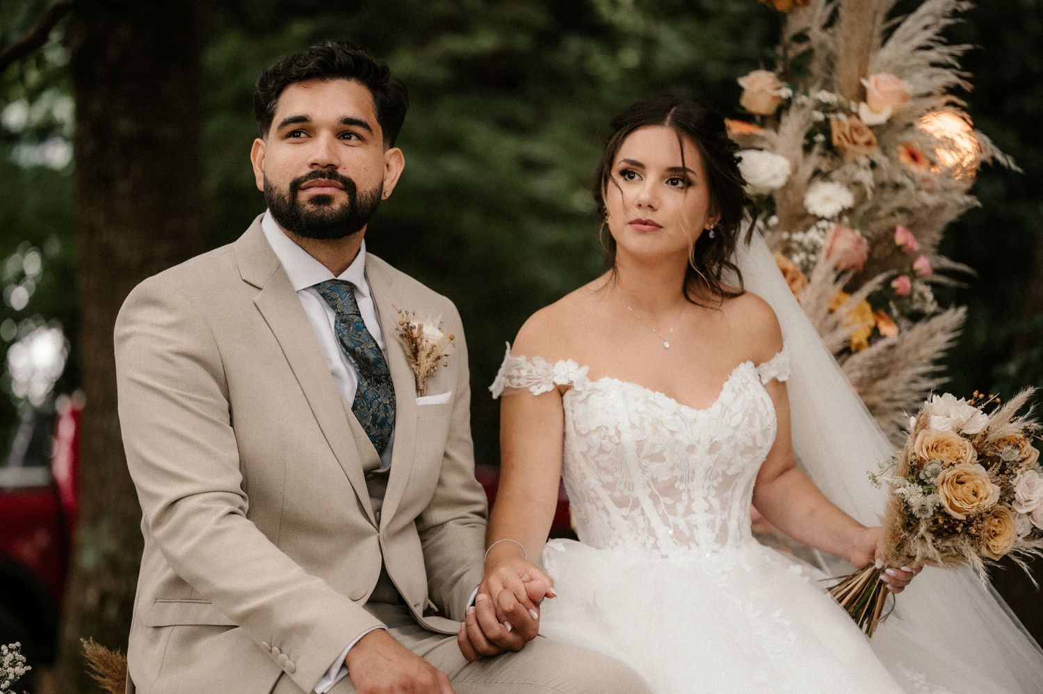 A wedding couple seated together during an outdoor ceremony with dried floral decorations.