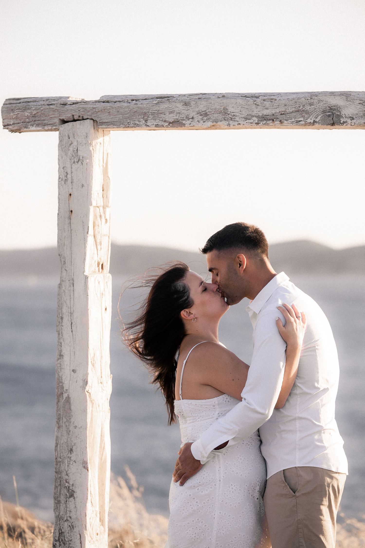Couple sharing romantic kiss under wooden arch structure at sunset.