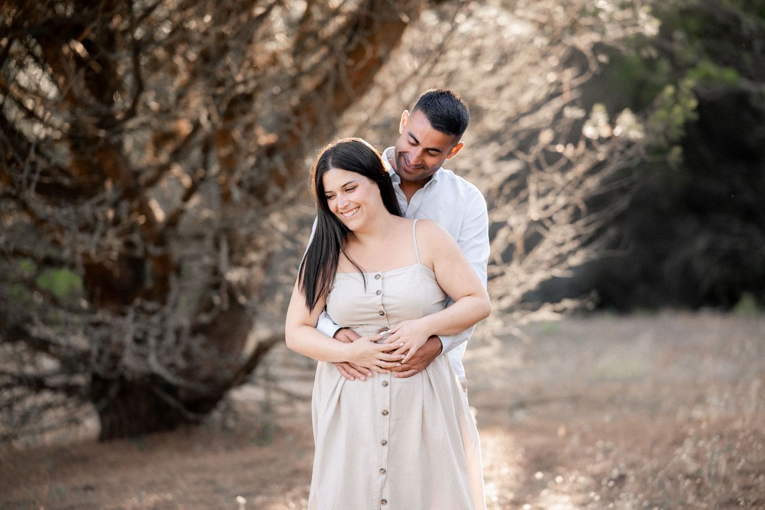 A couple in white outfits embraces during a romantic outdoor photoshoot in a natural desert setting.