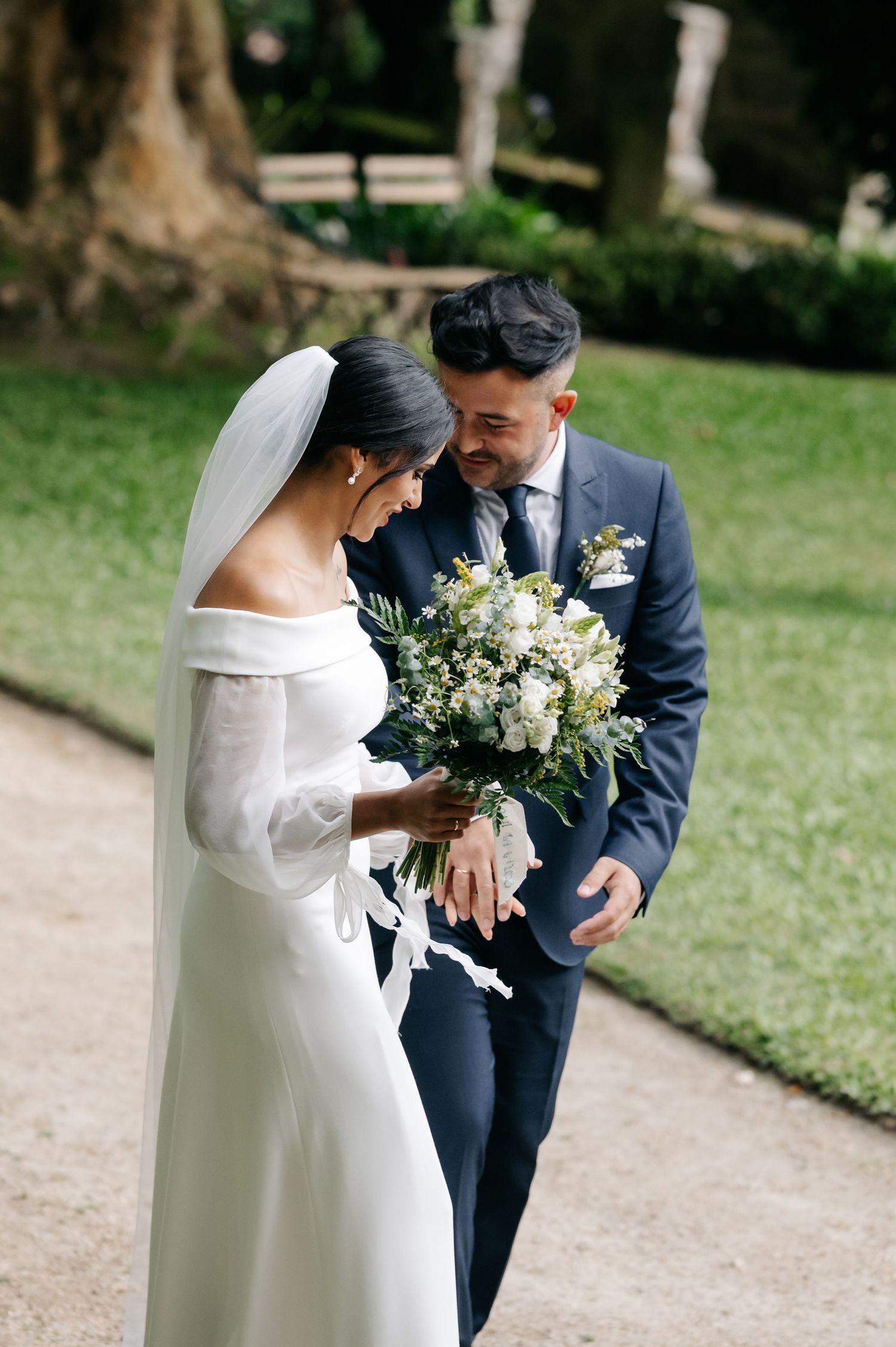 A bride in a white dress and groom in navy suit share intimate moments in a garden setting during their wedding photoshoot.
