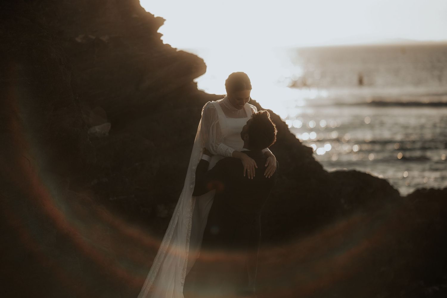A couple in wedding attire stands silhouetted against a dramatic sunset on the beach.