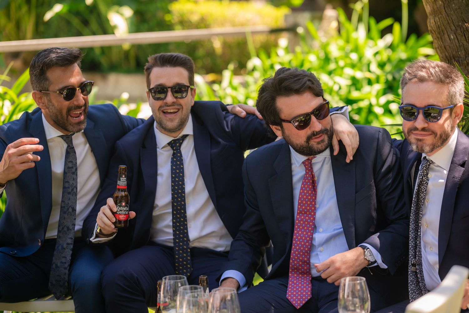 Group of formally dressed guests laughing and socializing at an outdoor event wearing sunglasses and suits.