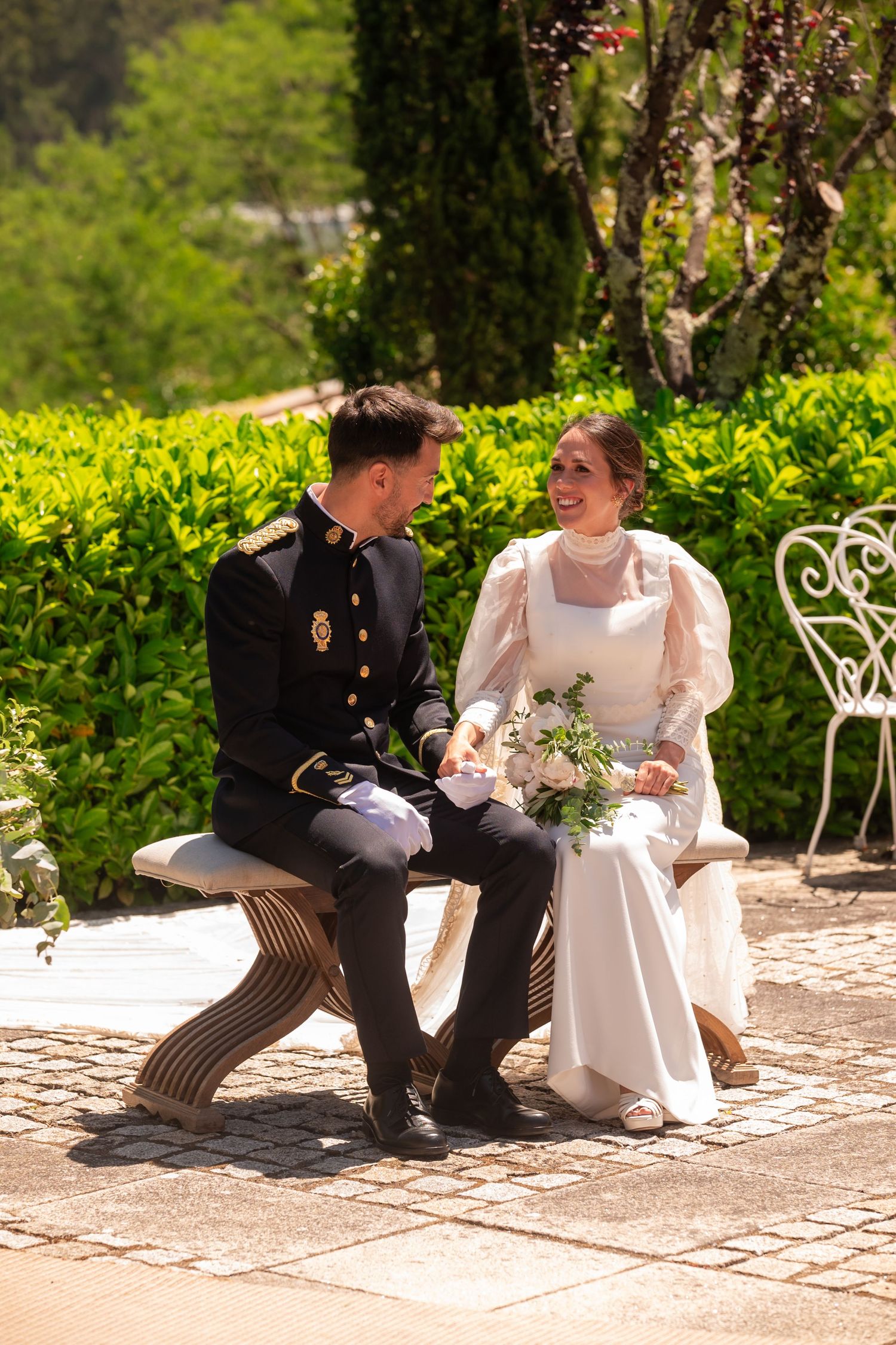 A military groom and bride in white dress share an intimate moment on a garden bench.