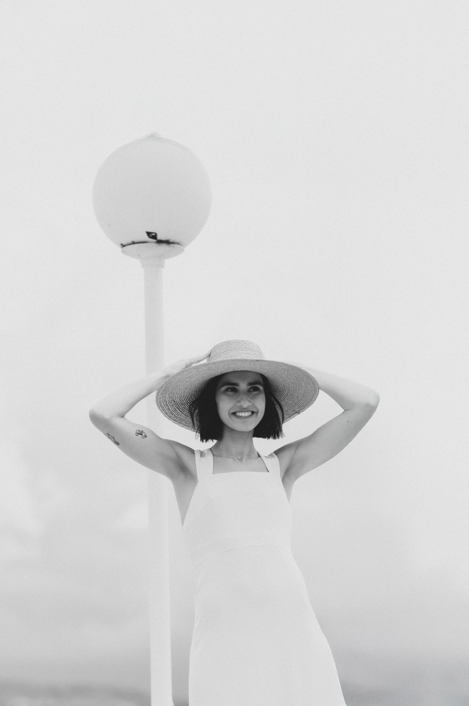 Artistic black and white portrait of bride in flowing dress and dramatic wide-brimmed hat against minimal background.