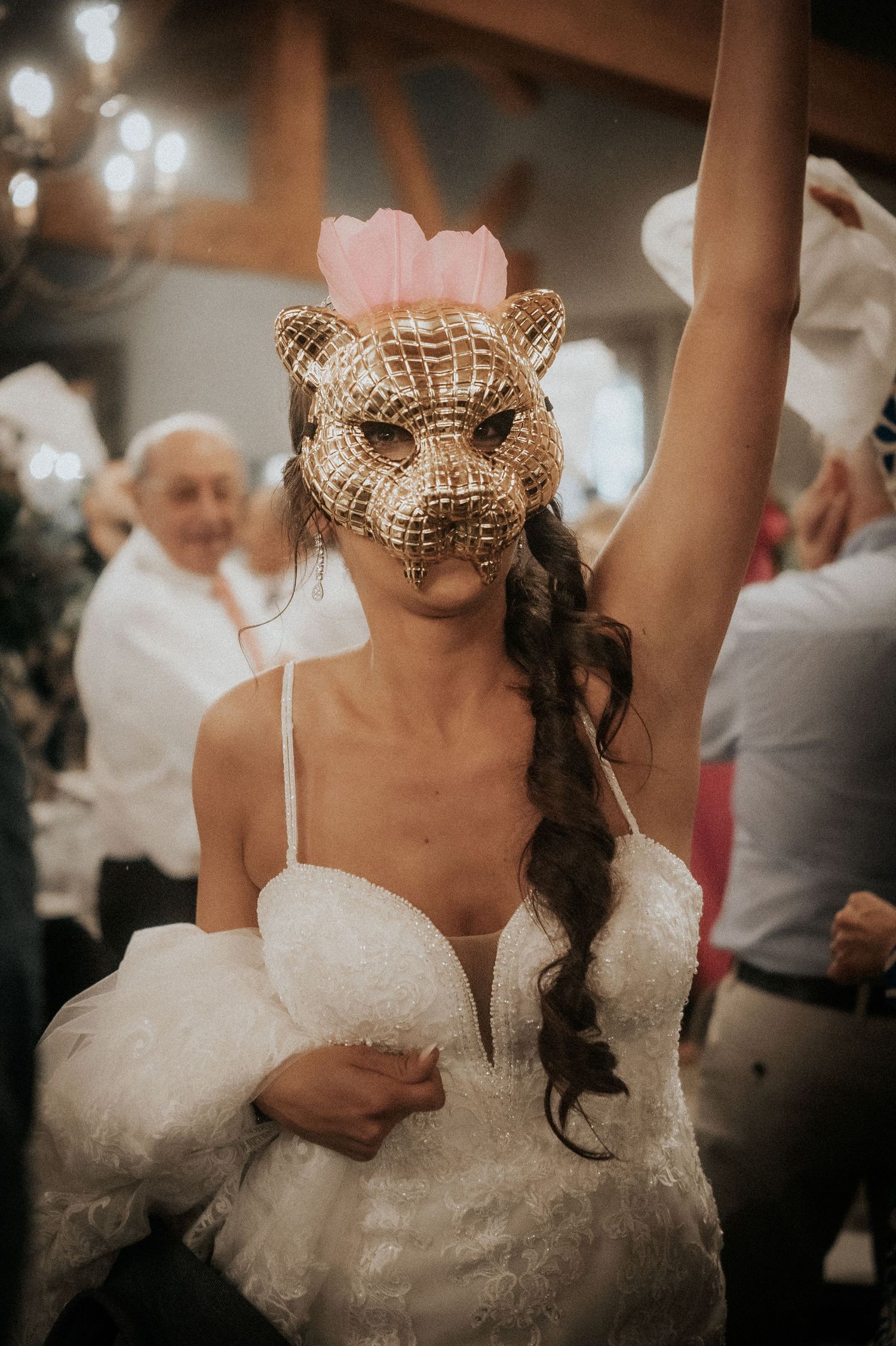 A person in a white wedding dress dances while wearing an ornate gold animal mask with a pink crown at a reception.